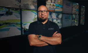 OnView executive standing inside the headquarters Live Surveillance Monitoring center with security camera feeds displayed behind him.