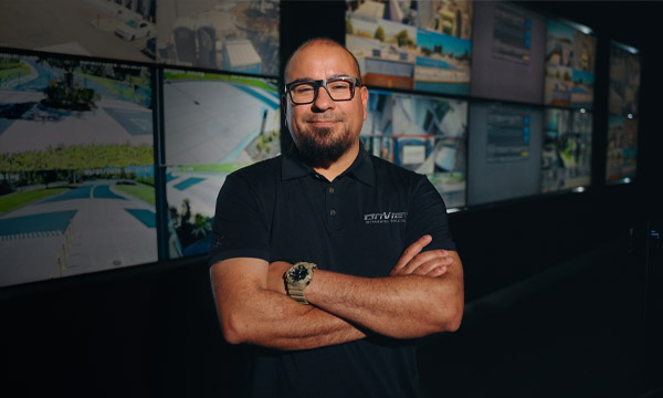 OnView executive standing inside the headquarters Live Surveillance Monitoring center with security camera feeds displayed behind him.